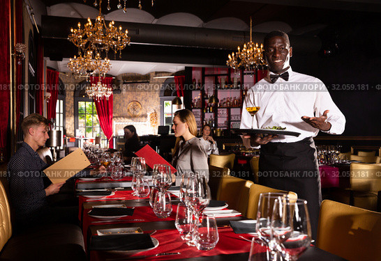 Hospitable african-american waiter standing with serving tray meeting restaurant guests. Стоковое фото, фотограф Яков Филимонов / Фотобанк Лори
