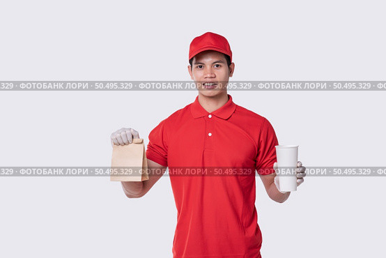 Image of a happy young delivery man in red cap blank t-shirt uniform standing with empty brown craft paper packet isolated on light gray background studio. Стоковое фото, агентство Ingram Publishing / Фотобанк Лори