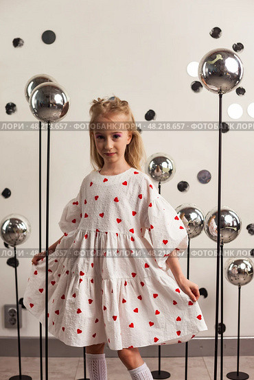 In a creative studio filled with playful decor, a 6-year-old girl gracefully twirls while wearing a charming white dress adorned with red hearts. Стоковое фото, фотограф Aleksandr Golubev / Фотобанк Лори