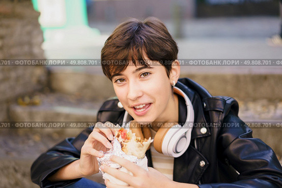 In a lively urban setting filled with energy and movement, a young person sporting a trendy short hairstyle and wearing headphones is happily sitting on the stairs while enjoying a delicious burrito,. A young person joyfully enjoys a delicious burrito while relaxing outdoors in the city. Стоковое фото, агентство Ingram Publishing / Фотобанк Лори