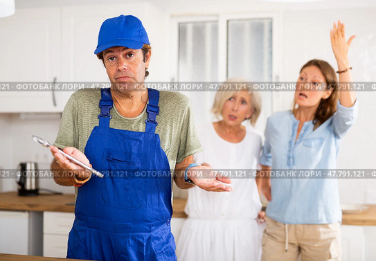 Inattentive plumber in uniform spreading his arms to the sides while being scolded by two women in the kitchen. Стоковое фото, фотограф Яков Филимонов / Фотобанк Лори