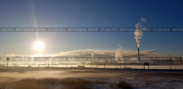Industrial landscape in winter against a backdrop of bright sunlight. Snow-covered and slightly frosted grass is visible in the foreground. In the distance are industrial chimneys from which thick white steam or smoke rises, creating a characteristic winter atmospheric effect. The sun is low above the horizon, illuminating the frame with golden rays and creating the feeling of a frosty morning. Steam, fog and smoke beautifully disperse in the air, emphasizing the contrast between nature and industrialization. The photo conveys the atmosphere of a cold day, industrial power and ecology. Стоковое фото, фотограф Анна Кривицкая / Фотобанк Лори