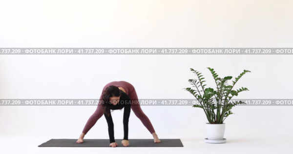 Инструктор йоги выполняет упражнение Паривритта Ардха Прасарита Падоттанасана, интенсивную позу растяжки, тренируется в леггинсах и коротком топе с длинными рукавами, стоя на коврике в студии. Стоковое видео, видеограф Сергей Чайко / Фотобанк Лори