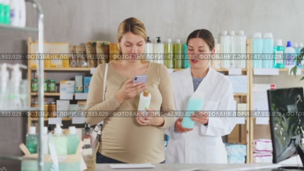 Interested pregnant woman using smartphone to check ingredients of organic shampoo at drugstore, while smiling professional female pharmacist in background suggesting different pregnancy-safe option. Стоковое видео, видеограф Яков Филимонов / Фотобанк Лори