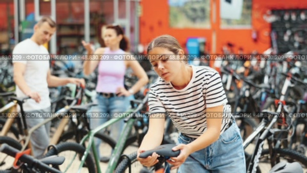 Interested young female cyclist choosing new bike for cycling trips in busy store, carefully picking comfortable bicycle saddle. Стоковое видео, видеограф Яков Филимонов / Фотобанк Лори