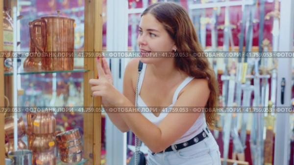 Interested young woman viewing various copper utensils placed on display case of souvenir store. Стоковое видео, видеограф Яков Филимонов / Фотобанк Лори