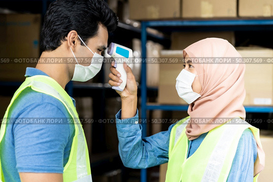 Islam asian warehouse worker taking temperature to worker before getting in factory after reopening from city lockdown from COVID-19 coronavirus pandemic in distribution center environment. Side view. Стоковое фото, агентство Ingram Publishing / Фотобанк Лори