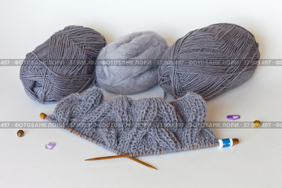 Knitting a warm gray scarf from wool and mohair yarn with bulky knitted braids. The row counter is worn on a knitting needle. Skeins of yarn on a white background. Winter handicraft and hobby. Стоковое фото, фотограф Виктория Катьянова / Фотобанк Лори