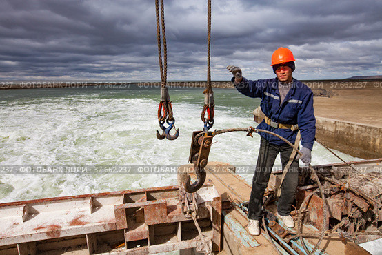 Kok-aral, Kazakhstan - May 03, 2012: Small Aral Sea Kok-aral dam. Worker lifts water shutter weighting agent. Steel rope and crane hooks. Showing hand signals to crane operator. Редакционное фото, фотограф Alexey Rezvykh / Фотобанк Лори