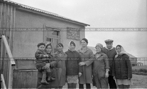 Коллективный фотопортрет людей позирующих стоя рядом с деревенским домом, одна из женщин держит ребёнка на руках, сельские жители, Лорино, Чукотка, 1970 год, колхоз Ленина. Редакционное фото, фотограф Варфоломей Тетерин / Фотобанк Лори