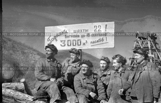 Коллективный портрет Шахтёров во время перерыва, мужчины позируют стоя рядом с плакатом с обращением к горнякам, деревянная конструкции позади них, Эмельджак, Якутия, 1961. Редакционное фото, фотограф Варфоломей Тетерин / Фотобанк Лори