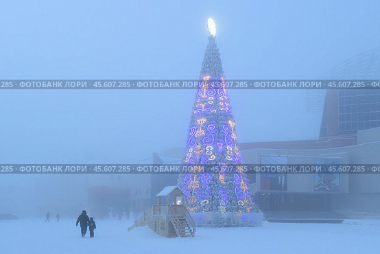 Красивая новогодняя ель в Якутске. Зимний городской пейзаж. Мимо рождественской ёлки идёт мужчина с ребёнком. Площадь Орджоникидзе, Город Якутск, Республика Саха (Якутия), Сибирь, Россия. Большая искусственная ёлка, украшенная традиционными якутскими орнаментами. Праздничное новогоднее настроение. Холодная погода и морозный туман. (2024 год). Редакционное фото, фотограф Андрей Степанов / Фотобанк Лори