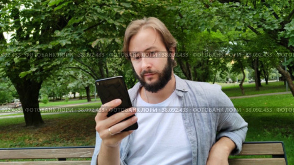 Красивый мужчина общается через мессенджер смартфона, сидя на скамейке в городском общественном парке. Бородатый парень читает новости, или устанавливает приложение, или пишет сообщение в своем мобильном телефоне. Портрет, крупный план. Handsome caucasian man communicates via smartphone messenger while sitting on bench in city public park. Bearded guy read news, or install app, or write message in his mobile phone. Portrait, close-up. Стоковое видео, видеограф Ирина / Фотобанк Лори