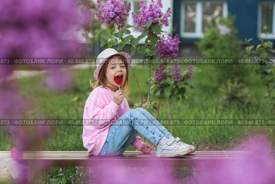 Lifestyle. A beautiful little girl in pink clothes with a lollipop in the shape of a heart. The girl licks the candy. The concept of love and sweets. Стоковое фото, фотограф Photojuli / Фотобанк Лори