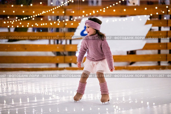 Little girl in a pink sweater and skirt is skating on a winter evening on an outdoor ice rink lit with garlands. Стоковое фото, фотограф Мария Мороз / Фотобанк Лори