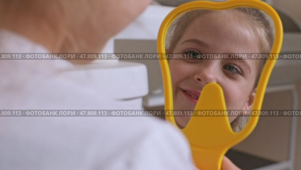 Lovely little girl looking in the mirror at her teeth after teeth surgery in a pediatric stomatology. A little girl sits in a dental chair and examines her cured teeth while looking in the mirror. Стоковое видео, видеограф Ирина Михайличенко / Фотобанк Лори