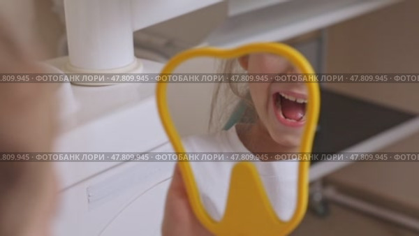 Lovely little girl looking in the mirror at her teeth after teeth surgery in a pediatric stomatology. A little girl sits in a dental chair and examines her cured teeth while looking in the mirror. Стоковое видео, видеограф Ирина Михайличенко / Фотобанк Лори