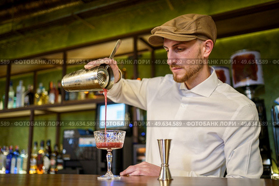 Low angle of bearded man in white shirt and cap filling glass goblet with fruit cocktail while working in bar in evening. Barman pouring cocktail into glass. Стоковое фото, агентство Ingram Publishing / Фотобанк Лори