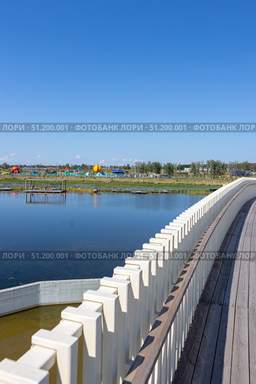 Magnitogorsk, Russian Federation - August 24, 2025: Scenic urban resort attraction featuring a wooden walkway beside a tranquil lake, colorful playgrounds in the background, ideal for family recreation and leisure activities. Редакционное фото, фотограф Ирина Ручушкина / Фотобанк Лори