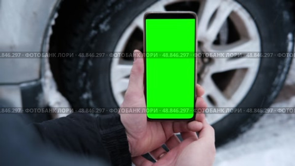 Male hands holding a smartphone with a bright green screen against the background of a car wheel. Automotive technology. Mobile applications for drivers. Road safety. Using gadgets. Chroma key. Стоковое видео, видеограф Евгений Демидюк / Фотобанк Лори