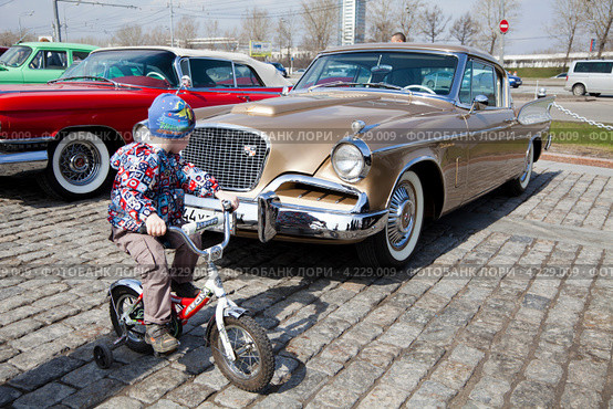Маленький мальчик проезжает на велосипеде мимо автомобиля Studebaker Golden Hawk на ралли классических автомобилей клуба РККА на Поклонной горе в Москве, Россия (2012 год). Редакционное фото, фотограф Николай Винокуров / Фотобанк Лори