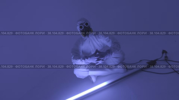Man in chemical protection suite sitting in front of fluorescent lamp. Стоковое видео, видеограф Aleksey Kidisyuk / Фотобанк Лори