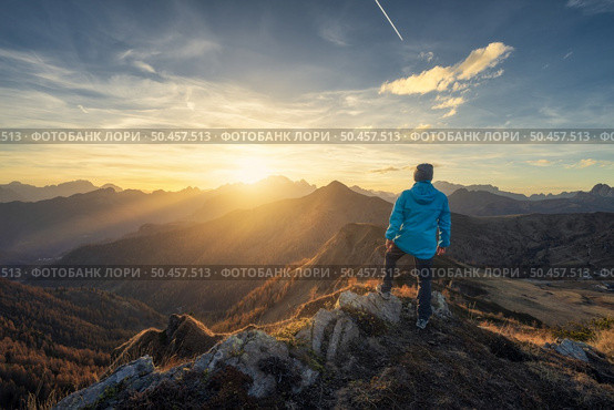 Man on stone on the hill and beautiful mountains in haze at colorful sunset in autumn. Dolomites, Italy. Sporty guy, mountain ridges in fog, orange grass and trees, blue sky with sun in fall. Hiking. Man on stone on the hill and beautiful mountains in haze. Стоковое фото, агентство Ingram Publishing / Фотобанк Лори