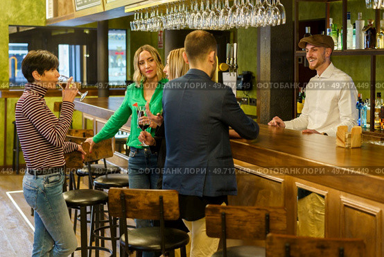 Man ordering some beverages from barkeeper while standing near happy talking women on weekend day in pub. Happy men and women chatting in a beautiful bar. Стоковое фото, агентство Ingram Publishing / Фотобанк Лори
