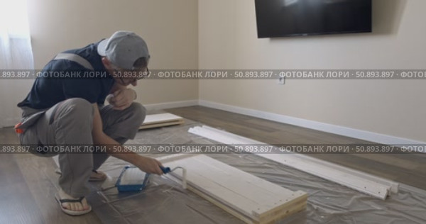 Man Professional carpenter painting wooden boards with white paint using roller brush in modern interior room during home renovation construction. Painting of wooden shelving. furniture assembly. Стоковое видео, видеограф Сергей Тимофеев / Фотобанк Лори