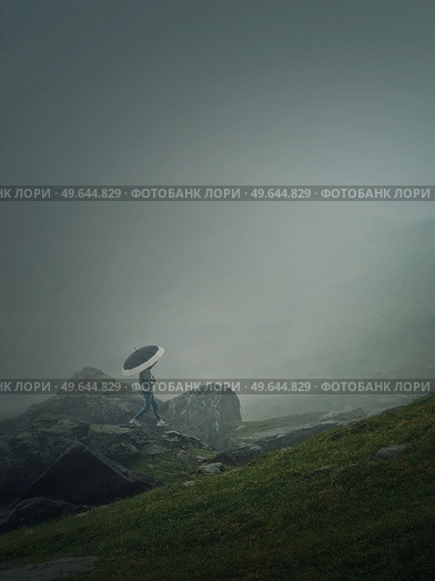 Man with umbrella climbing a mountain hill in a gloomy weather with dense mist. Moody hiking scene with a wanderer walking on the cliffs of the foggy valley. Стоковое фото, агентство Ingram Publishing / Фотобанк Лори