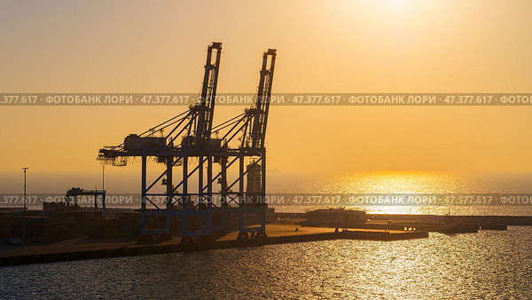 Manama, Bahrain - Jan 08 2024, Panoramic view of Bahrain commercial port with blue cranes, buildings and containers, with sea on foreground, by the morning, dawn, sunrise, Manama, Bahrain. Стоковое фото, фотограф Сергей Богомяко / Фотобанк Лори