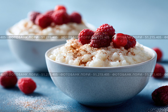 Milky Rice pudding with superberries and red raspberry sauce, served in a white bowl on blue tablecloth background. Rice porridge topped with some raspberries and cinnamon sprinkled. Old English style. Стоковое AI-изображение, AI-художник Марина Демидюк / Фотобанк Лори