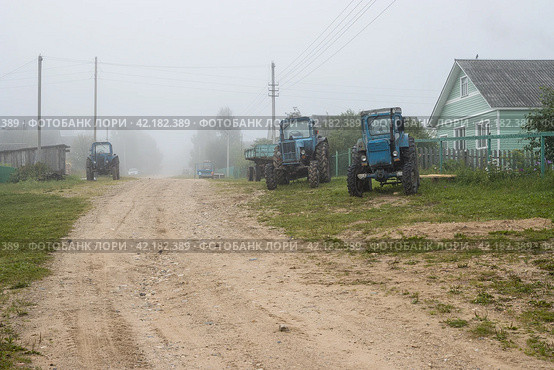 Misty morning in the countryside. View of a deserted village street. Old tractors on the side of a dirt road. Foggy rural landscape. Wooden houses behind fences. Travel across Russia. Vologda region. Стоковое фото, фотограф Андрей Степанов / Фотобанк Лори