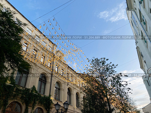 Moscow, Russia, July 22, 2025: view of Kuznetsky Most Street. Festive decoration lamps hang overhead historical street. Charming architecture features intricate details with greenery climbing. Редакционное фото, фотограф Olga Monako / Фотобанк Лори
