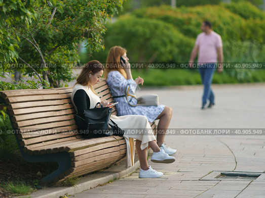 Moscow, Russia - May 20, 2021: Young pretty women sitting and resting on wooden bench in the public park. Hot spring day. Everybody use smartphones. Youth leisure concept. Walking man in distance. Редакционное фото, фотограф YuryKara / Фотобанк Лори