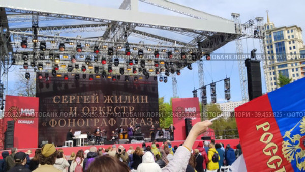 МОСКВА, РОССИЯ - 9 мая 2022 года: Люди в городском парке смотрят концерт, посвященный Дню Победы 9 мая. Женщина размахивает российским флагом. Выступление Сергея Жилина и оркестра "Фонограф Джаз". MOSCOW, RUSSIA - MAY 9, 2022: People in the city park watch a concert dedicated to Victory Day on May 9. A woman waves a Russian flag. Performance by Sergey Zhilin and the Phonograph Jazz Orchestra. Редакционное видео, видеограф Ирина / Фотобанк Лори