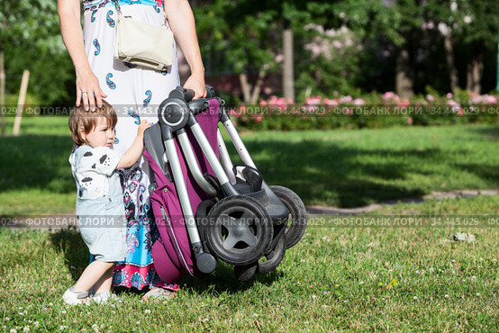 Mother calm down her toddler daughter while standing with pushchair in other hand, summer green lawn, copyspace. Стоковое фото, фотограф Кекяляйнен Андрей / Фотобанк Лори