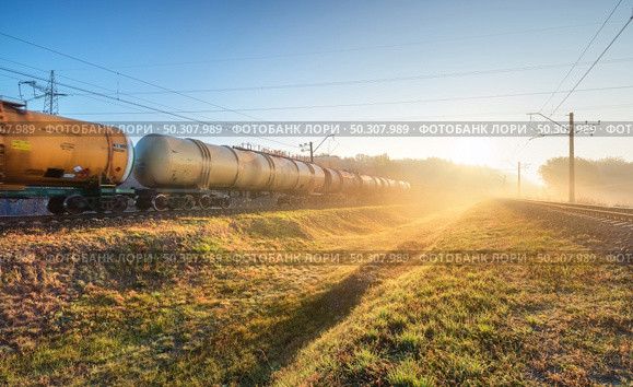 Moving freight train in fog at sunrise in autumn. Industrial landscape with train in motion, railroad, green grass, trees, gold sunbeams, foggy trees, blue sky in fall. Railway station. Transportation. Moving freight train in fog at sunrise in autumn. Industrial. Стоковое фото, агентство Ingram Publishing / Фотобанк Лори