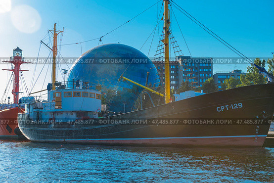 Museum ship SRT-129, moored at the embankment next to the Planetarium of the Museum of the World Ocean in Kaliningrad. It is an old fishing trawler with a black hull and yellow masts, carefully restored for museum purposes. In the background is the spectacular globe-shaped planetarium ball and modern buildings. To the right are reflections of sunlight on the water. To the left of the ship, a bright red lighthouse ship is partially visible. The scene is full of marine and scientific symbolism, and the day is clear and sunny. (2020 год). Редакционное фото, фотограф Анна Кривицкая / Фотобанк Лори