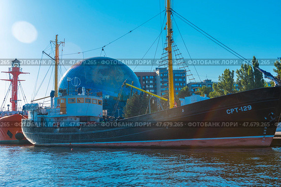 Museum ship SRT-129, moored at the embankment next to the Planetarium of the Museum of the World Ocean in Kaliningrad. It is an old fishing trawler with a black hull and yellow masts, carefully restored for museum purposes. In the background is the spectacular globe-shaped planetarium ball and modern buildings. To the right are reflections of sunlight on the water. To the left of the ship, a bright red lighthouse ship is partially visible. The scene is full of marine and scientific symbolism, and the day is clear and sunny. (2020 год). Редакционное фото, фотограф Анна Кривицкая / Фотобанк Лори