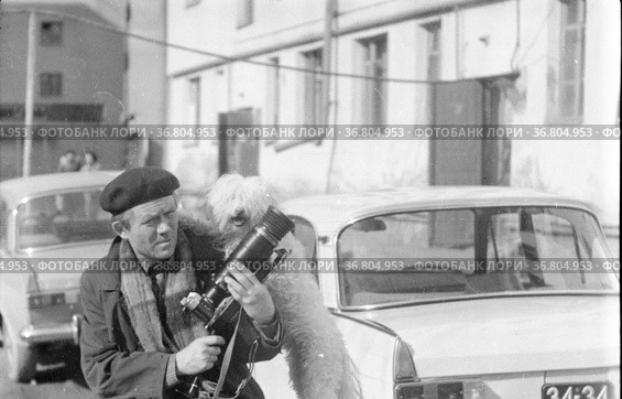 Небольшая собака рядом с фотографом, мужчина готовит камеру для фотосъемки, он во дворе дома, пес позирует оперившись на мужчину, белая болонка на двух лапах , Тетерин, Фатпулов, Долли, Якутия, май 1980 года. Редакционное фото, фотограф Варфоломей Тетерин / Фотобанк Лори