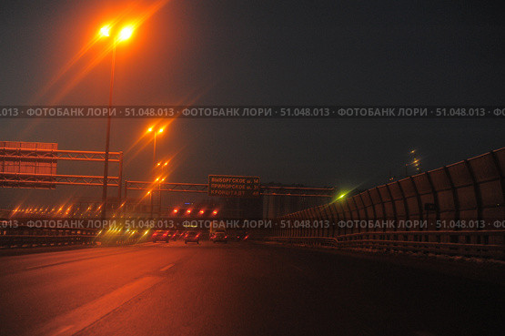 Night view of the Saint Petersburg Ring Road, illuminated by bright street lamps with a warm orange light. The road surface sparkles in the light of headlights and lanterns, creating an atmosphere of movement in the evening city. In the foreground are several cars moving in the indicated directions: Vyborgskoye Highway, Primorskoye Highway, Kronstadt. High noise protection screens are installed on the right, and information signs and signs are above the road. The scene conveys a sense of urban dynamics, high-speed traffic and infrastructure of a megalopolis in the dark. Стоковое фото, фотограф Анна Кривицкая / Фотобанк Лори