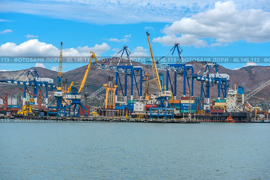 Novorossiysk Commercial Sea Port, one of the largest ports in Russia, located on the Black Sea coast. The frame shows cargo ships, port cranes, container yards and part of the industrial infrastructure. In the background are the mountain ranges surrounding the city.Numerous blue and yellow gantry cranes used for loading and unloading ships.Cargo containers stored in the port area.Commercial ships at the berths undergoing cargo operations.Tugboats used to maneuver large ships.Mountains, characteristic of the Novorossiysk landscape.Bright blue sky with clouds, creating a contrast with the industrial landscape. (2023 год). Редакционное фото, фотограф Анна Кривицкая / Фотобанк Лори