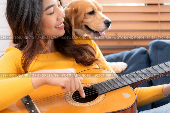 On a weekend, A young Asian woman and her beagle puppy are playing the guitar and singing cheerfully together in the living room of a house. Стоковое фото, агентство Ingram Publishing / Фотобанк Лори