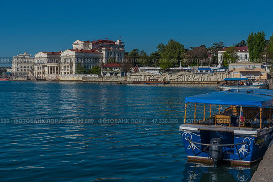 One of the most recognizable views of Sevastopol is the central embankment of the city with a view of historical buildings, snow-white architecture and a smooth sea surface. On the right, a pleasure boat with a blue awning is moored, ready for a sea excursion. In the background are majestic buildings with columns, one of which is the building of the Historical Boulevard and the Black Sea Fleet Museum, as well as other cultural and administrative buildings.The bright sun, blue sky and calm water create an atmosphere of southern comfort and resort tranquility. Sevastopol appears as a city with a rich history, a maritime soul and a promising future. (2021 год). Редакционное фото, фотограф Анна Кривицкая / Фотобанк Лори