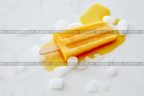 Orange melting ice cream lolly with ice slices on a gray marble table with a copy of the space. The concept of summer dessert.Flat lay. Orange fruit ice cream on a stick with slices of ice on a gray marble background with space for text. Flat lay. Стоковое фото, агентство Ingram Publishing / Фотобанк Лори