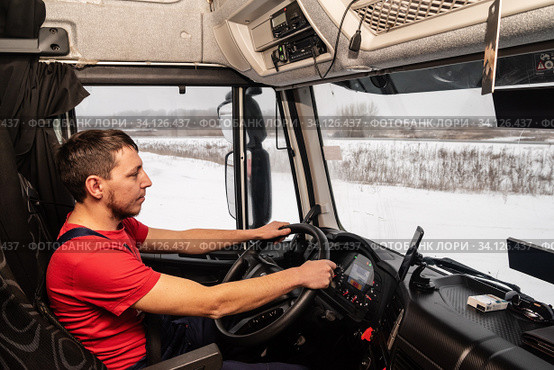Orenburg, Russia - December, 17, 2019: Driver in a red T-shirt at the workplace at the wheel of a truck. Inside view of the cab. Редакционное фото, фотограф Вадим Орлов / Фотобанк Лори