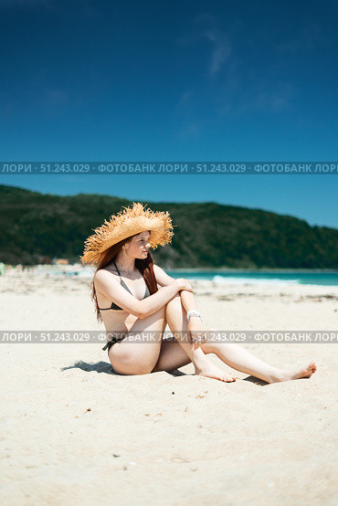 Peaceful summer scene: young woman in straw hat sits barefoot on sand, looking thoughtfully toward sea moment of solitude and connection with nature. Стоковое фото, фотограф Енваев Андрей / Фотобанк Лори