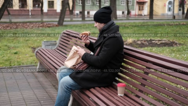 Pensive hipster drinking coffee sitting on a bench in the urban park. Handsome man wearing black coat eating street food, hot dog with paper cup in hand in the city. Stylish bearded middle-aged male. Стоковое видео, видеограф Марина Демидюк / Фотобанк Лори