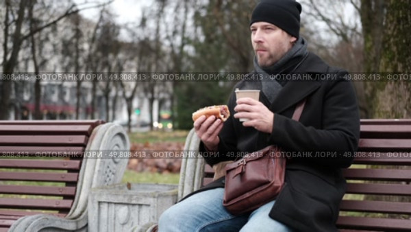 Pensive hipster drinking coffee sitting on a bench in the urban park. Handsome man wearing black coat eating street food, hot dog with paper cup in hand in the city. Stylish bearded middle-aged male. Стоковое видео, видеограф Марина Демидюк / Фотобанк Лори